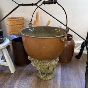 May include: A round copper pot with a metallic finish, featuring a curved iron handle with a dark wooden grip. The pot is secured by metal rivets on the sides and rests upon a textured, weathered stone pedestal with an ornate, carved base.