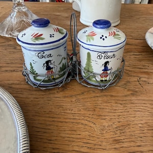 May include: A pair of ceramic spice jars with lids, held in a wire basket. The jars are white with blue lids and feature hand-painted designs of a woman, trees, and the words "Sea" and "Flour".