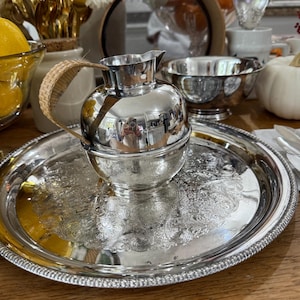 Puede incluir: Una jarra plateada con un asa tejida se encuentra sobre una bandeja plateada a juego con un diseño floral ornamentado. La jarra y la bandeja reflejan los objetos circundantes, incluyendo un cuenco y un espejo. La escena está sobre una superficie de madera.