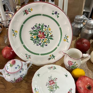 Puede incluir: Un juego de platos de cerámica blanca con un diseño floral en tonos de rosa, verde, amarillo y morado. Los platos tienen un borde festoneado y un borde rojo. También hay una jarra de crema y un azucarero a juego.