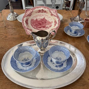 May include: A silver-plated oval serving tray holding a silver creamer and two blue and white transferware teacups with matching saucers. Behind the tray sits a scalloped-edge plate featuring a red and white landscape scene with a castle.