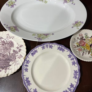 May include: Three white ceramic plates with floral designs. The largest plate is oval shaped with purple and white flowers. The two smaller plates are round, one with a purple floral design and the other with a multi-colored floral design.
