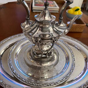 May include: A silver tea set, including a teapot with a curved handle and a matching oval tray. The teapot sits on a stand, and the tray has an ornate, embossed border. The set is reflective and appears to be made of metal.