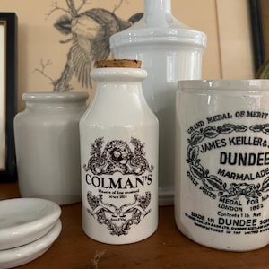 Pode incluir: Três frascos de cerâmica branca com tampas de cortiça. O frasco do meio é rotulado "COLMAN'S Masters of fine mustard since 1814". O frasco à direita é rotulado "GRAND MEDAL OF MERIT JAMES KEILLER & SON DUNDEE MARMALADE ONLY PRIZE MEDAL FOR MARMALADE LONDON 1863 Contents 1 lb. Net MADE IN DUNDEE SCOTLAND JAMES KEILLER & SON LTD. DUNDEE SCOTLAND MANUFACTURED IN THE UNITED KINGDOM."