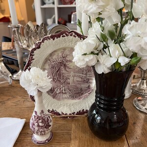 May include: Two ceramic vases with floral designs. The vase on the left is a small, white vase with a purple floral pattern. The vase on the right is a larger, black vase with a wide mouth. Both vases are filled with white carnations.