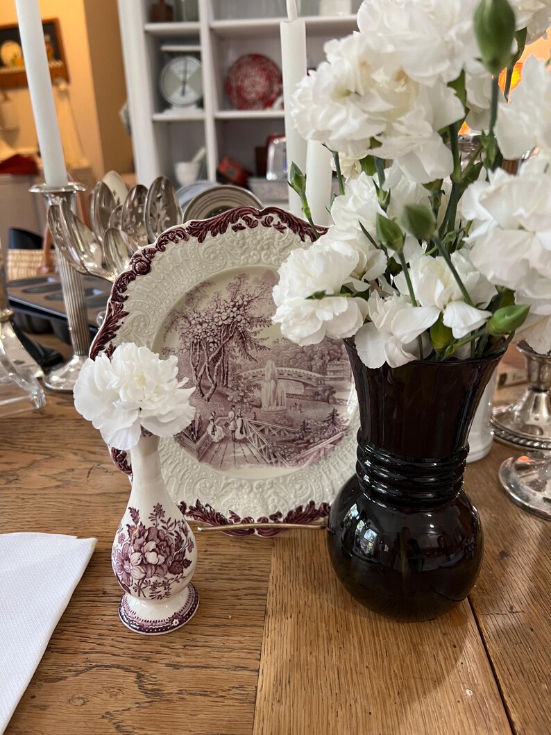 May include: Two ceramic vases with floral designs. The vase on the left is a small, white vase with a purple floral pattern. The vase on the right is a larger, black vase with a wide mouth. Both vases are filled with white carnations.