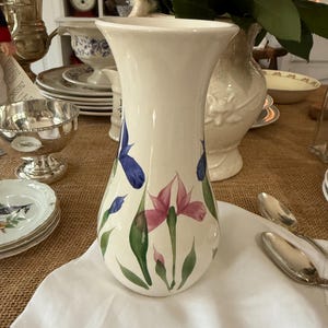 May include: A cream-coloured ceramic vase with a flared top, hand-painted with blue and pink iris flowers and green leaves. The vase is a decorative item, suitable for holding flowers and adding elegance to any room.