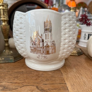 Puede incluir: Jarrón de cerámica color crema con una ilustración dorada de un edificio. El jarrón tiene un patrón de diamantes texturizado en los lados y una base de pedestal. El texto en el jarrón dice "The Cathedral, City of Bury."