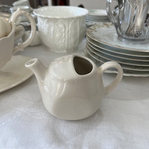 Op de afbeelding: Een witte keramische roomkan met een afgeronde vorm en een klein tuitje. De roomkan staat op een wit tafelkleed.