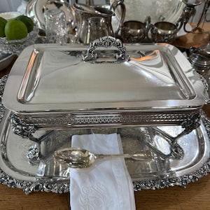May include: A rectangular silver-plated serving dish with a lid, featuring ornate floral handles and feet. The dish rests on a matching silver tray with an intricate embossed border. A silver spoon lies on a white linen napkin with embroidered initials.