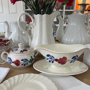 Puede incluir: Tetera, jarra de crema, azucarero y salsera de cerámica blanca con un diseño floral azul y rojo. El azucarero y la salsera tienen una tapa. La tetera y la jarra de crema tienen un asa. La salsera tiene un platillo a juego. Un plato blanco está en primer plano.