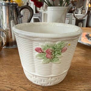 May include: Off-white ceramic basket-weave planter with a floral design. The planter features a textured basket-weave pattern and a raised relief of pink flowers and green leaves. The planter is sitting on a wooden table.