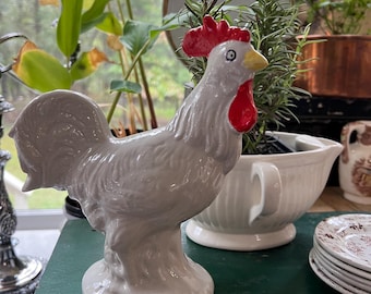 Pode incluir: Uma figura de galo de cerâmica branca com crista e barbela vermelhas. O galo está em pé sobre uma superfície verde.