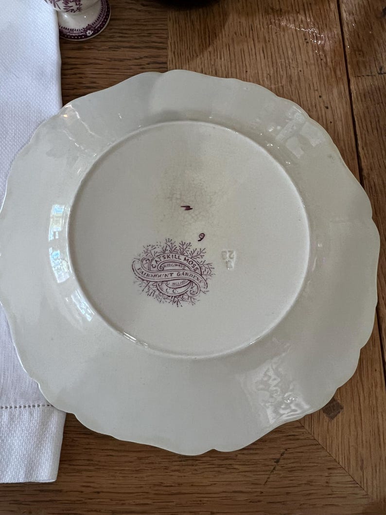 May include: A white ceramic plate with a scalloped edge. The back of the plate has a purple and white design with the text "Samuel Moss, Original, Patent Gardens, Miller & Co."
