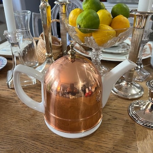 May include: A copper and white teapot with a lid and a white handle. The teapot is sitting on a wooden table with silver candlesticks and a bowl of lemons and limes.