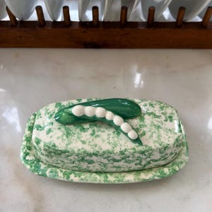 Può includere: Un porta burro in ceramica verde e bianco con un coperchio a forma di baccello di piselli. Il coperchio è decorato con un baccello di piselli verde con piselli bianchi.