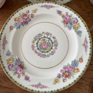 May include: White porcelain plates with a floral design. The plates feature a gold rim and a border of green and gold detailing. The floral pattern includes pink, purple, yellow, and blue flowers. A smaller floral arrangement is in the center.