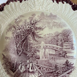 May include: A vintage ceramic plate with a raised, scalloped edge and a purple floral design. The center of the plate features a detailed illustration of a park scene with a bridge, fountain, and people walking.
