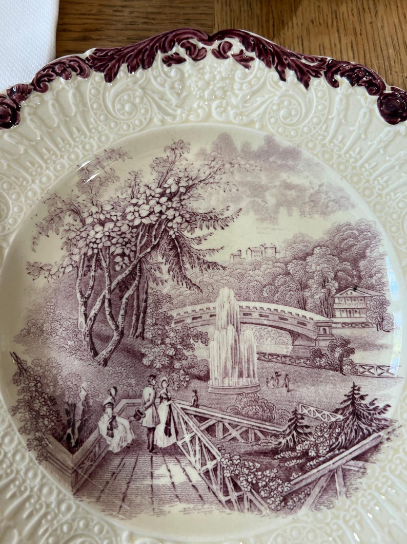 May include: A vintage ceramic plate with a raised, scalloped edge and a purple floral design. The center of the plate features a detailed illustration of a park scene with a bridge, fountain, and people walking.
