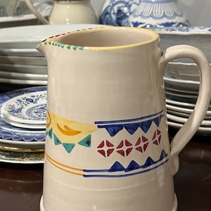 May include: A white ceramic pitcher with a colorful geometric design. The pitcher has a wide mouth and a handle. The design features yellow, orange, red, blue, and green colors.
