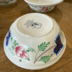 May include: White ceramic bowls with floral designs in pink, blue, and green. The bowls have a classic shape and are likely part of a dinnerware set. The underside of one bowl shows a stamp with text.