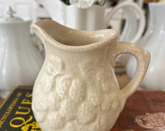 Könnte beinhalten: Ein kleiner, weißer Keramik-Milchkännchen mit einer strukturierten, craquelé-Glasur. Der Milchkännchen hat ein erhabenes Design aus Trauben und Blättern.