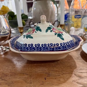 Pode incluir: Uma travessa de cerâmica creme com tampa. A tampa apresenta uma pequena figura no topo e é decorada com padrões florais vermelhos e verdes. A travessa tem uma borda azul estampada. A travessa está sobre uma superfície de madeira.