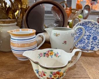 Könnte beinhalten: Eine Sammlung von Vintage-Sahnekännchen und Servierschalen auf einem Holztisch. Die Kännchen weisen Blumen- und Streifenmuster in verschiedenen Farben auf, darunter Gelb, Blau und Orange. Ein Teller mit blau-weißem Muster ist ebenfalls zu sehen.