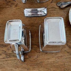 May include: A collection of polished silver serving pieces, including a large bowl, two small rectangular dishes, and other utensils. The dishes have the text "Fairpoint" and "TPNS C329" engraved on them. The items are arranged on a wooden surface.