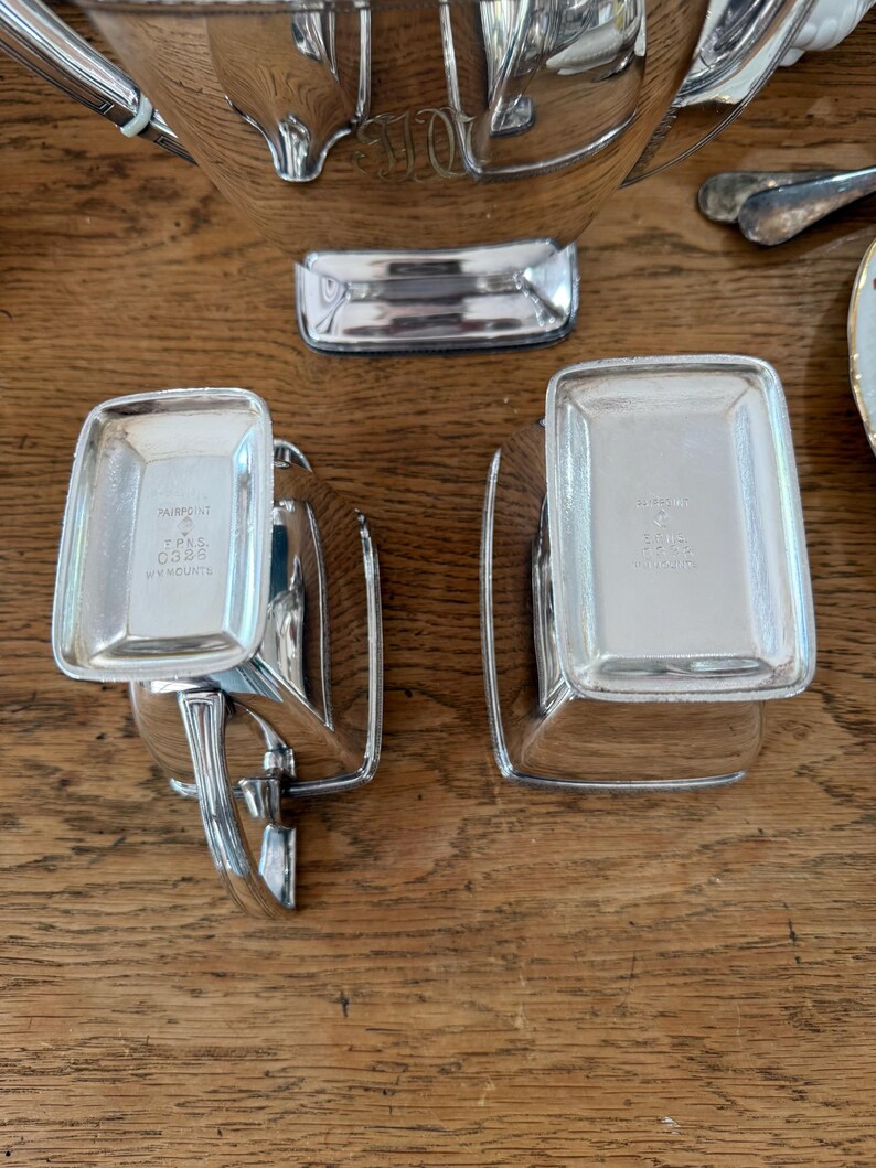 May include: A collection of polished silver serving pieces, including a large bowl, two small rectangular dishes, and other utensils. The dishes have the text "Fairpoint" and "TPNS C329" engraved on them. The items are arranged on a wooden surface.