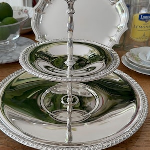 Puede incluir: Una bandeja plateada de dos niveles con un asa decorativa. Las bandejas tienen un borde con cuentas y una superficie reflectante. El asa es adornada y conecta los dos niveles. Otras bandejas y botellas están en el fondo.