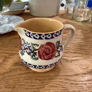 Puede incluir: Jarra pequeña de cerámica con cuerpo crema e interior beige. Decorada con una rosa roja y detalles florales azules y verdes. Un patrón geométrico azul bordea la parte superior e inferior.