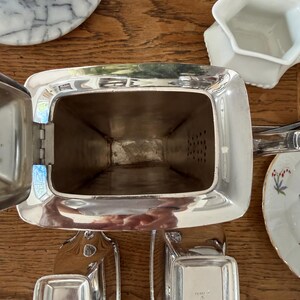 May include: A silver-plated tea set, including a rectangular teapot with a hinged lid and two small rectangular trays. The teapot has a handle and a spout. The set is displayed on a wooden surface.