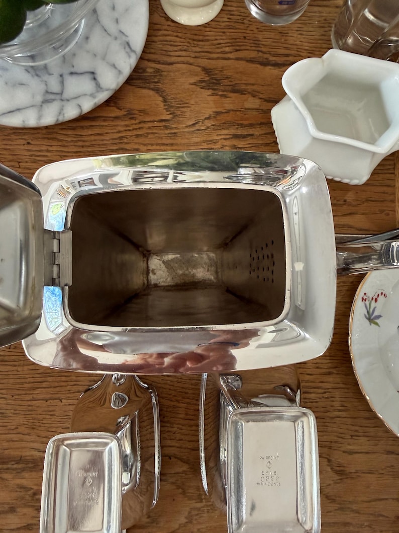 May include: A silver-plated tea set, including a rectangular teapot with a hinged lid and two small rectangular trays. The teapot has a handle and a spout. The set is displayed on a wooden surface.