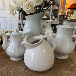 May include: A set of four white ceramic jugs, including a large jug with a wide mouth, a smaller jug with a spout, and two smaller jugs with handles. The jugs are arranged on a brown hessian surface.