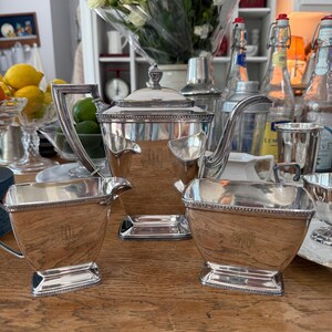 May include: A silver tea set with a teapot, creamer, and sugar bowl. The set has a rectangular shape with ornate handles and trim. The teapot has a lid with a decorative finial. The set is displayed on a wooden surface.
