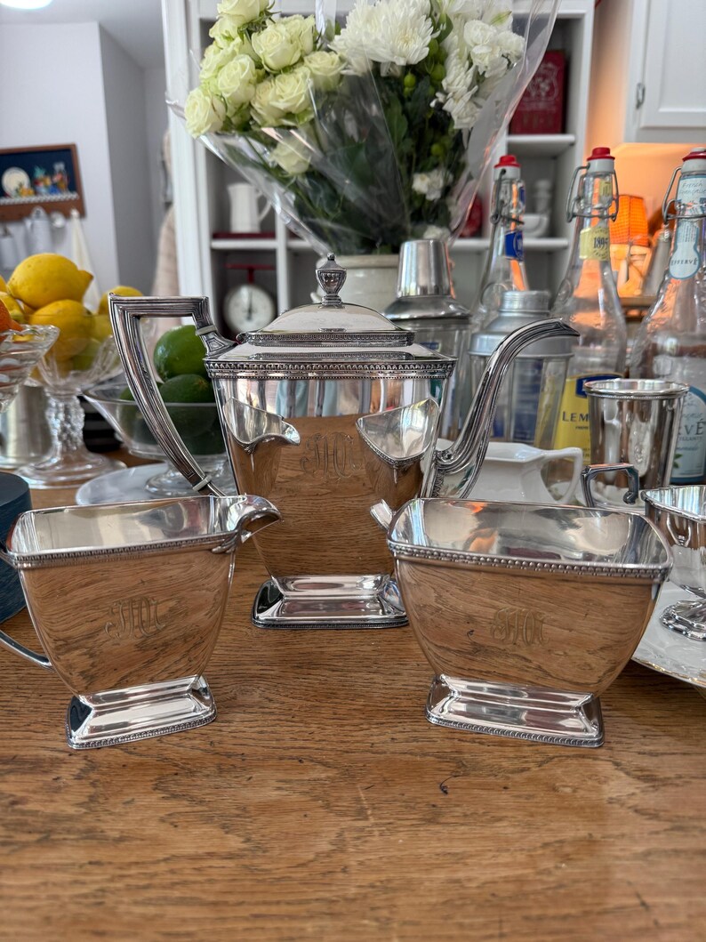 May include: A silver tea set with a teapot, creamer, and sugar bowl. The set has a rectangular shape with ornate handles and trim. The teapot has a lid with a decorative finial. The set is displayed on a wooden surface.