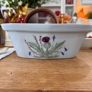 May include: A rectangular, off-white ceramic dish with a floral thistle design. The illustration features a purple thistle flower, green leaves, and blue bellflowers. The dish is likely for serving or baking.