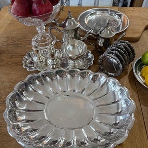 May include: A collection of silver serving pieces, including a large scalloped platter, small teapots, a sugar bowl, and a salt and pepper set. A glass bowl holds several pomegranates. The items are arranged on a wooden table.