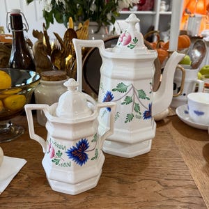 May include: A white ceramic teapot and creamer set with a floral design featuring pink and blue flowers and green leaves. The teapot has a tall, angular shape with a handle and spout. The creamer is smaller, with a similar design.