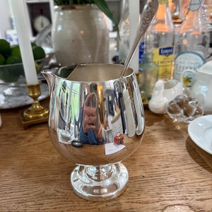 May include: A shiny silver creamer with a small spout and a pedestal base. A silver spoon rests inside the creamer. The creamer reflects the surrounding objects, including a person taking the photo. The creamer is on a wooden table.