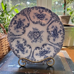Könnte beinhalten: Ein weißer Keramikteller mit einem blauen und weißen Blumenmuster. Der Teller hat einen gewellten Rand und wird auf einem goldenen Metallständer präsentiert.