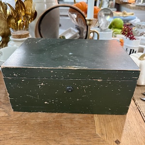 May include: A dark green wooden box with a slightly distressed finish. The box has a rectangular shape with a hinged lid and a small, round lock. The box is sitting on a wooden table.