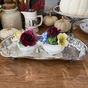 May include: Two white porcelain flower-shaped containers with colorful floral arrangements sit on a silver tray. The flowers include red, yellow, and blue blooms. The tray is on a wooden surface, with pumpkins and other decorative items in the background.
