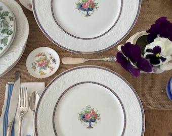 May include: Two white ceramic dinner plates with blue and gold trim and a floral design in the center. The plates are on a brown burlap surface.