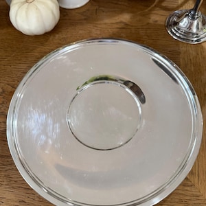 May include: A circular, silver-coloured serving tray with a raised edge and a smaller, circular indentation in the centre. The tray is reflective and sits on a wooden surface. Decorative pumpkins and a mug are in the background.