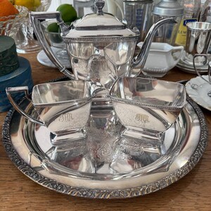 May include: A silver tea set with a teapot, two cups, and a serving tray. The set has ornate detailing and is placed on a wooden surface. The cups and teapot have the initials "MH" engraved on them.