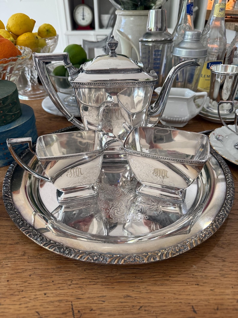 May include: A silver tea set with a teapot, two cups, and a serving tray. The set has ornate detailing and is placed on a wooden surface. The cups and teapot have the initials "MH" engraved on them.