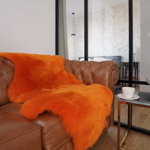 May include: A vibrant orange sheepskin rug draped over a brown leather tufted sofa. The rug features a thick, plush texture and a natural, irregular shape, providing a bold color contrast against the smooth, button-tufted leather upholstery.