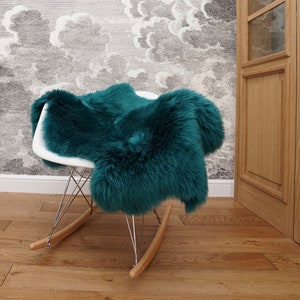 May include: A deep teal, long-haired sheepskin rug draped over a white molded plastic rocking chair. The chair features a white seat shell, a chrome wire base, and light-toned wooden rocker runners, creating a contrast with the dark fur.